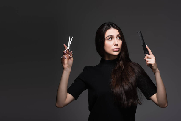 young woman with long shiny hair holding scissors and comb isolated on dark grey - Foto, afbeelding