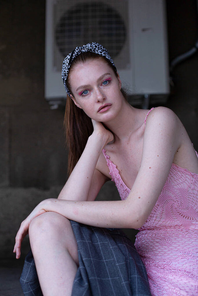Sitting white blond young woman with a ponytail in pink summer dress with blue eyes in an industrial area - Foto, Imagem