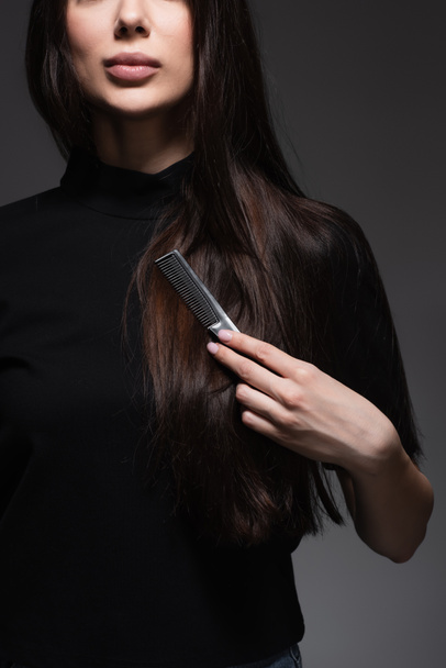 vista parziale di giovane donna spazzolatura lunghi capelli lucidi con pettine isolato su grigio scuro - Foto, immagini