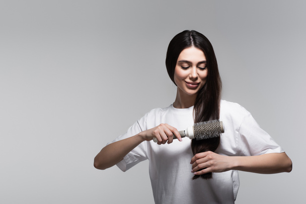 довольная женщина расчесывает гладкие длинные волосы тепловой щеткой, изолированной на сером - Фото, изображение