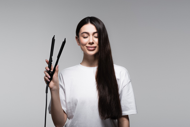 pleased young brunette woman holding hair straightener isolated on grey - Fotó, kép