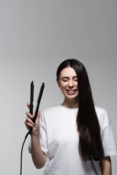 happy brunette woman holding hair straightener isolated on grey - Fotó, kép