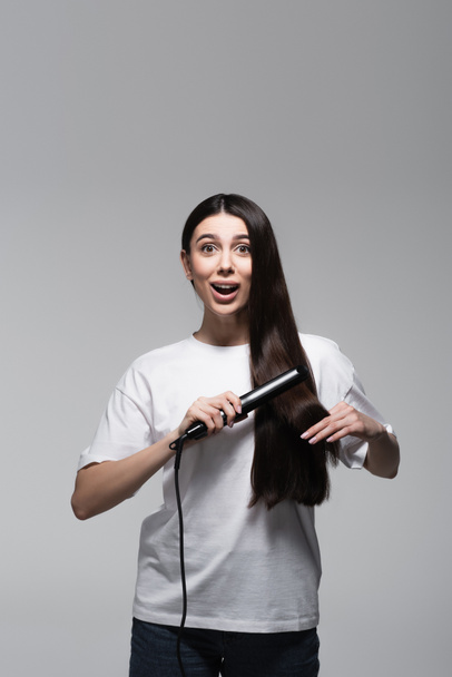 young amazed woman using hair straightener isolated on grey - Фото, изображение