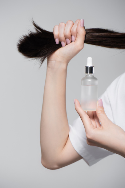 partial view of young woman holding bottle with oil and shiny hair isolated on grey - Foto, Imagem