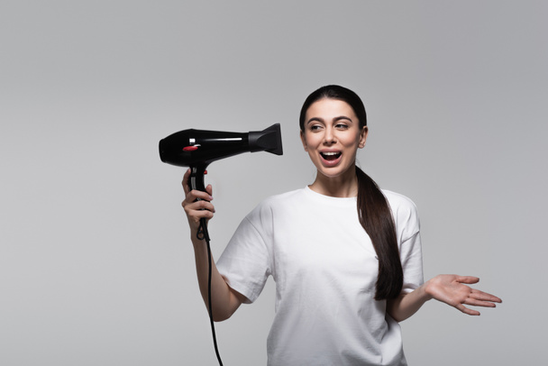 excited woman in white t-shirt holding hair dryer isolated on grey - Valokuva, kuva