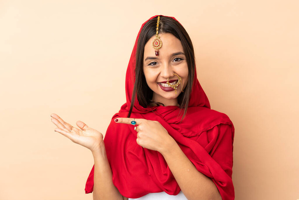 Молода індійська жінка, ізольована на бежевому тлі, тримає на долоні копіювальний простір уявної на долоні, щоб вставити оголошення. - Фото, зображення