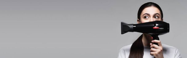 Nuori nainen valkoinen t-paita kattaa kasvot hiustenkuivaaja eristetty harmaa, banneri - Valokuva, kuva