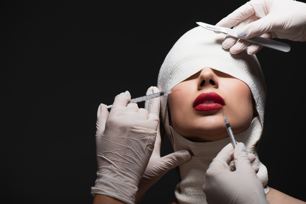 молодая женщина в эластичной повязке рядом с пластическими хирургами с хирургическим ножом и шприцами, изолированными на сером - Фото, изображение