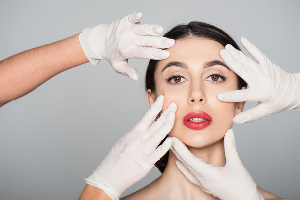 plastic surgeons in latex gloves touching face of brunette woman isolated on grey - Fotoğraf, Görsel