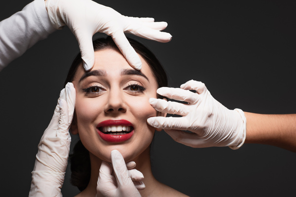 chirurdzy plastyczni w lateksowych rękawiczkach dotykający twarzy uśmiechniętej młodej kobiety odizolowanej na czarno  - Zdjęcie, obraz