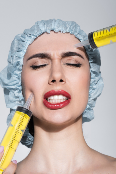 огромные шприцы возле лица напуганной молодой женщины в хирургической шапке, изолированной на сером - Фото, изображение