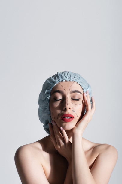 jeune femme en bonnet chirurgical touchant le visage avec des lignes marquées isolées sur gris - Photo, image