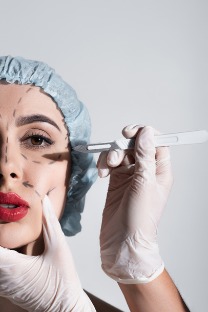 partial view of plastic surgeon in latex gloves holding scalpel near woman with marked lines on face isolated on grey - Photo, Image