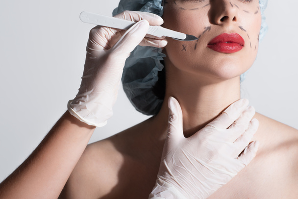 女性の赤い唇の近くにメスを保持するラテックス手袋の整形外科医のクロップドビュー顔にグレーに隔離されたマークラインと - 写真・画像