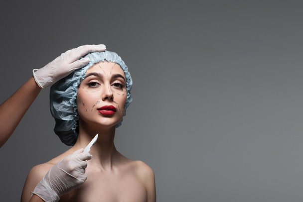 plastic surgeon in latex gloves holding scalpel near woman with marked lines on face and bare shoulders isolated on dark grey - Photo, Image