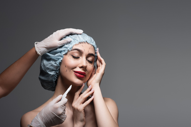 plastic surgeon holding scalpel near pleased woman in surgical cap with marked lines on face isolated on dark grey - Foto, immagini