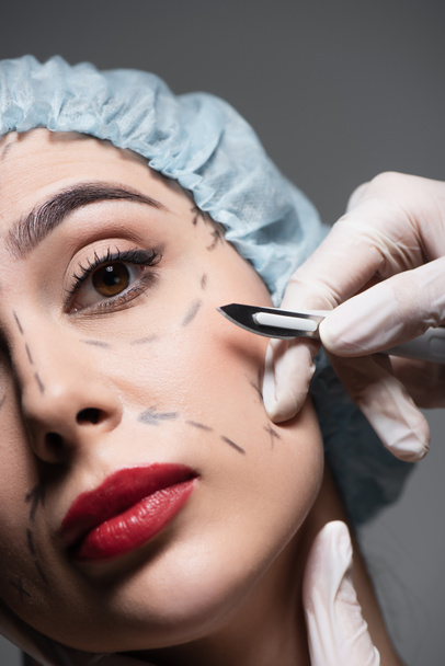 close up de cirurgião plástico em luvas de látex segurando bisturi perto de mulher com e linhas marcadas no rosto isolado em cinza - Foto, Imagem