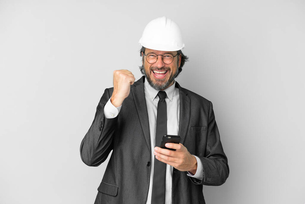 Jeune architecte avec casque sur fond isolé avec téléphone en position de victoire - Photo, image