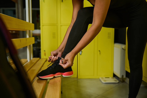 Close-up van een onherkenbare sportvrouw die schoenveters knoopt en haar voet op een houten bank zet in een kleedkamer van de sportschool - Foto, afbeelding