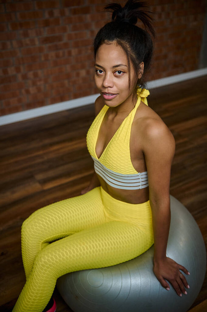 Magas látószögű kilátás egy vonzó afrikai sportnő, karcsú test sportoló fényes sárga melegítőben ül egy szürke fitness labda ellen piros tégla fal háttér fitness stúdió - Fotó, kép
