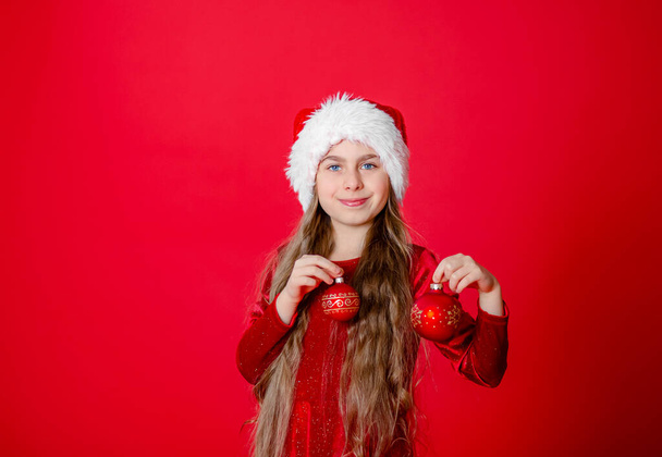 Noel Baba şapkası ve Noel topları takmış neşeli, parlak kırmızı bir arka planda izole edilmiş komik bir kız portresi. Çocuk eliyle işaret eder, metin için yer gösterir.. - Fotoğraf, Görsel