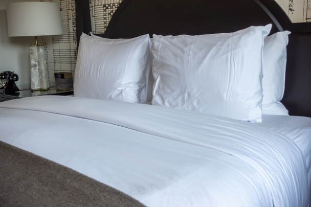 Vista en ángulo de una cama de hotel limpiamente hecha, con ropa de cama blanca y gris fresca y un cabecero negro - Foto, Imagen