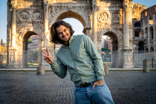 Feliz momento en Roma. Joven hombre sonriente posando para una foto haciendo el signo V delante del arco de Tito. - Foto, Imagen