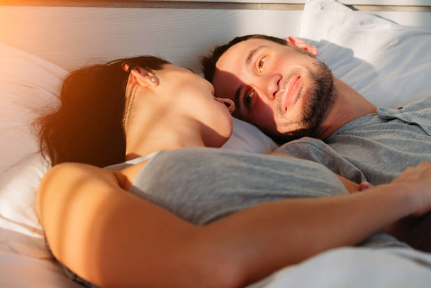 Jonge getrouwde echtparen worden 's morgens wakker in de slaapkamer en kijken elkaar aan. De man kijkt met een liefdevolle blik naar zijn jonge vrouw. Gelukkig familie goedemorgen concept. - Foto, afbeelding