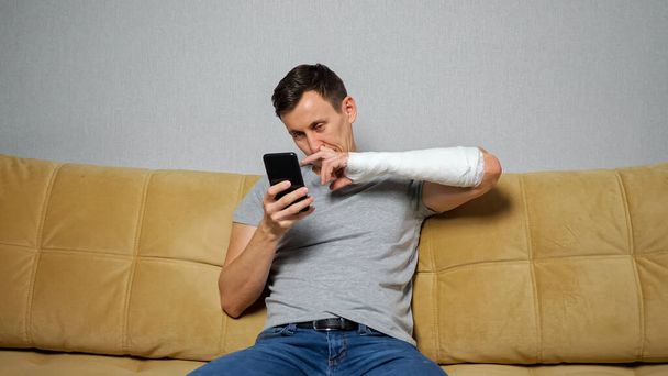 Brunet człowiek dłubie w nosie ze złamanym ramieniem przewijanie mediów społecznościowych - Zdjęcie, obraz