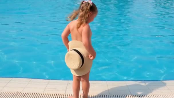 kleines Mädchen steht und bedeckt sich mit einem Sonnenhut vor dem Hintergrund des blauen Wassers des Pools oder Meeres. Sommerurlaub, Sonnencreme, Reisen. Selektiver Fokus auf Hut. - Filmmaterial, Video