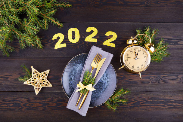 Festive table setting. Happy New Year 2022. Plate, silverware, fir branches, golden alarm clock.  - Photo, image