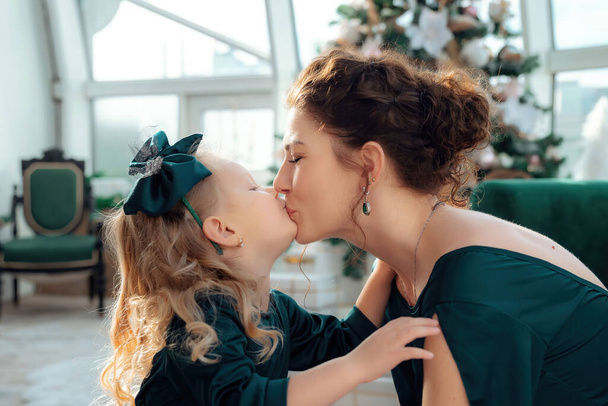 A mom kisses her little cute daughter. Caucasian mother and child dressed in festive green dresses. Xmas tree on the background. Christmas time is here - time for family. Close up portrait. - Photo, Image