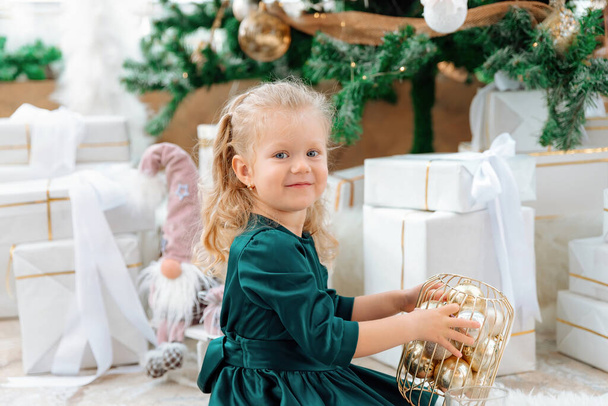 A three-year-old Caucasian girl in dress posing at a Christmas tree with a xmas balls in her hands. Child looks at the camera, smiles and happy. A lot of wrapped gift boxes on the background. - Фото, изображение