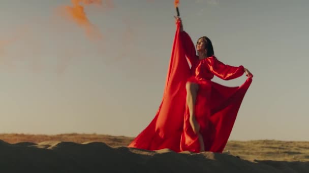 Uma mulher asiática em um vestido vermelho com uma bomba de fumaça laranja em suas mãos gira em dunas de areia - Filmagem, Vídeo