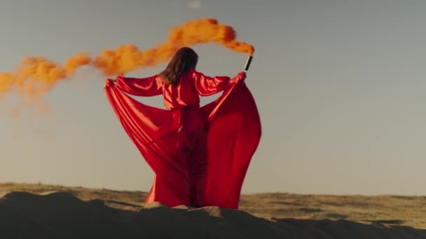 Una asiática vestida de rojo con una bomba de humo naranja en las manos gira sobre dunas de arena - Imágenes, Vídeo