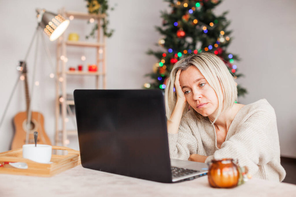 Frustrated woman in front of a laptop during the Christmas holidays. A disgruntled, upset middle-aged female at home on the background of a Christmas tree - Fotoğraf, Görsel