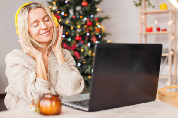 Happy middle-aged woman wearing headphones with a laptop on the background of a Christmas tree. Adult lady listens to music during the Christmas holidays - Fotoğraf, Görsel