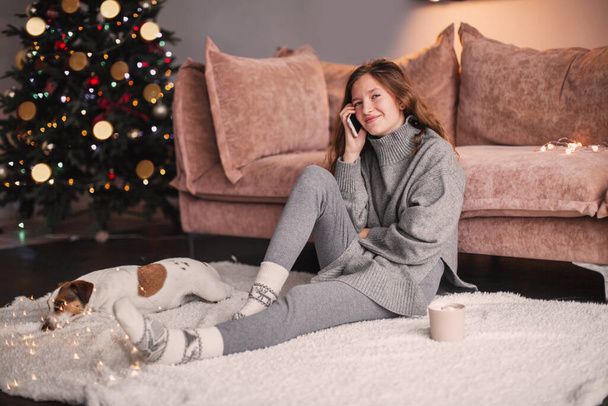 A jovem está a falar ao telefone em casa. Adolescente com celular está sentado em uma sala de estar contra uma árvore de Natal - Foto, Imagem