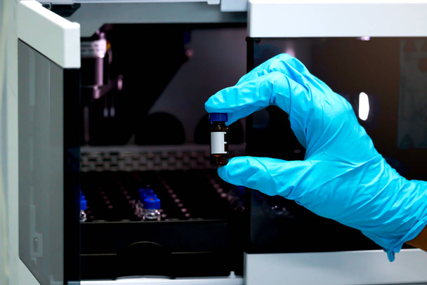 Naukowiec ręka z niebieskimi chemicznymi rękawicami ochronnymi trzymającymi fiolkę (mała butelka) do przeprowadzania eksperymentów. Chemik pracujący w laboratorium chemicznym z przyrządem HPLC (High Performance Liquid Chromatography). - Zdjęcie, obraz