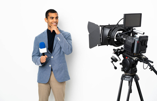Δημοσιογράφος Αφροαμερικάνος που κρατάει μικρόφωνο και μεταδίδει ειδήσεις σε απομονωμένο λευκό φόντο κοιτώντας στο πλάι και χαμογελώντας - Φωτογραφία, εικόνα