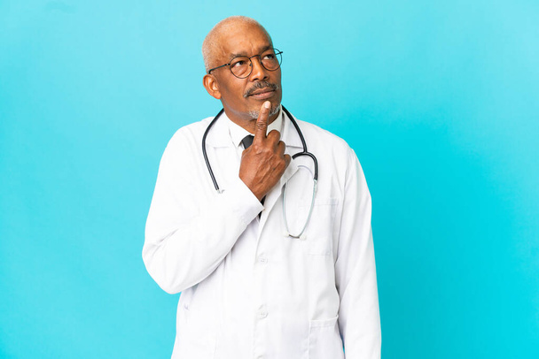 Senior arts man geïsoleerd op blauwe achtergrond met twijfels tijdens het omhoog kijken - Foto, afbeelding
