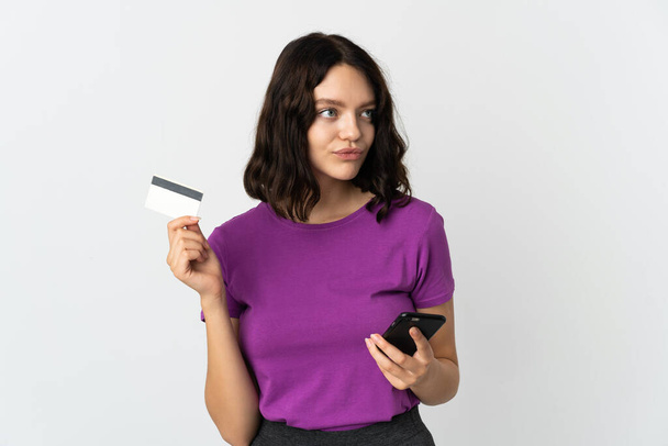 Teenager Ukrainian girl isolated on white background buying with the mobile with a credit card while thinking - Φωτογραφία, εικόνα