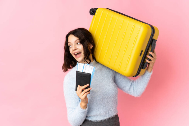 Підліткова українська дівчинка ізольована на рожевому фоні у відпустці з валізою та паспортом. - Фото, зображення