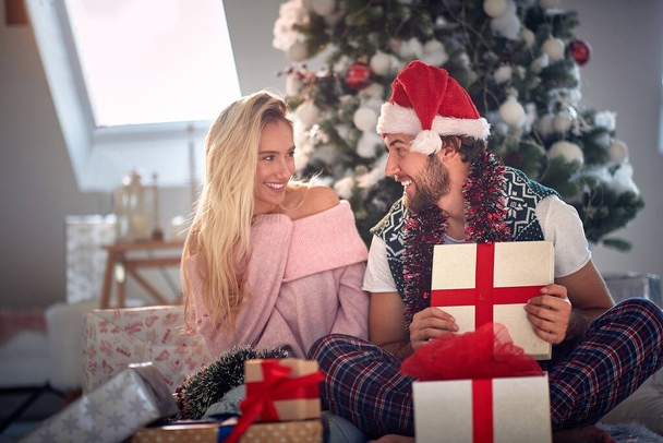 A guy is opening a christmas present he got from his girlfriend while they are sitting on the floor during christmas holidays in a festive atmosphere at home together.  - Photo, image