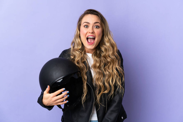 Mujer brasileña joven sosteniendo un casco de motocicleta aislado sobre fondo púrpura con expresión facial sorpresa - Foto, Imagen