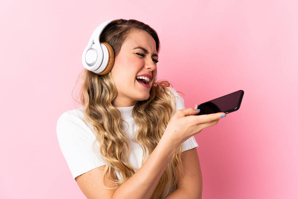 Νεαρή Βραζιλιάνα απομονωμένη σε ροζ φόντο ακούγοντας μουσική με κινητό και τραγουδώντας - Φωτογραφία, εικόνα