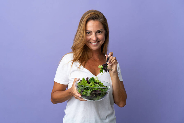 Mujer brasileña de mediana edad aislada sobre fondo púrpura sosteniendo un tazón de ensalada con expresión feliz - Foto, Imagen