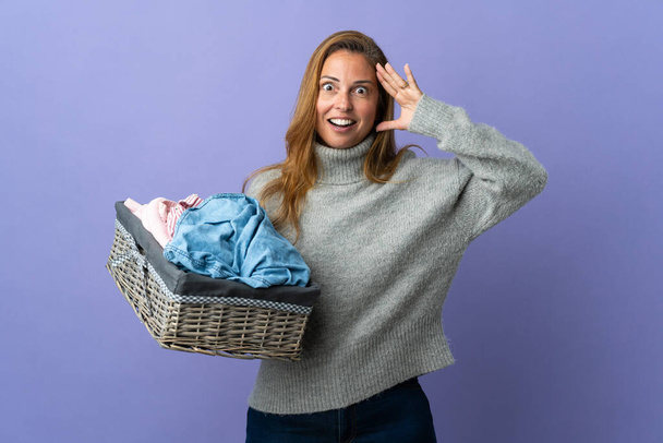 Frau mittleren Alters hält einen Wäschekorb mit Überraschungsausdruck auf lila Hintergrund - Foto, Bild