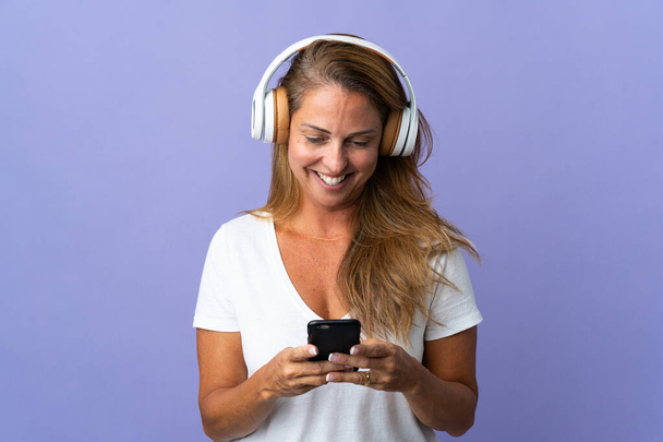 Középkorú brazil nő elszigetelt lila háttér hallgat zenét, és keresi a mobil - Fotó, kép