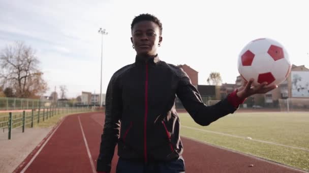 Egy fiatal afro-amerikai lány áll egy városi stadionban a sugárnapon, és egy focilabdát tart kinyújtott kezén. Lassú mozgás. - Felvétel, videó